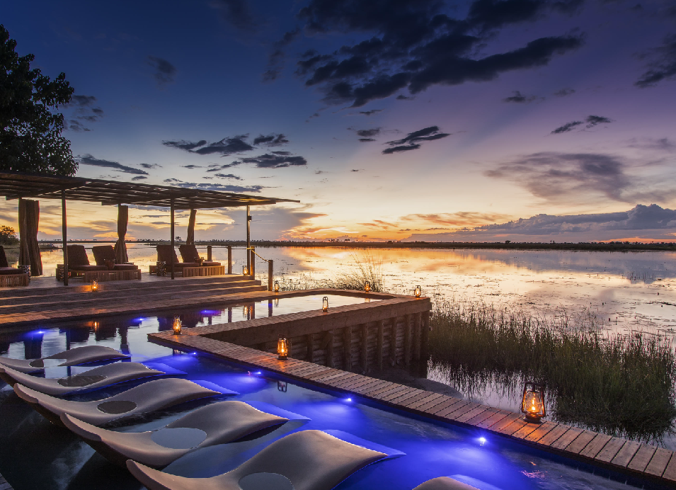 Luxury safari lodge pool overlooking a tranquil wetland at sunset in Botswana, with a wooden deck, lounge chairs, and open-air seating area.