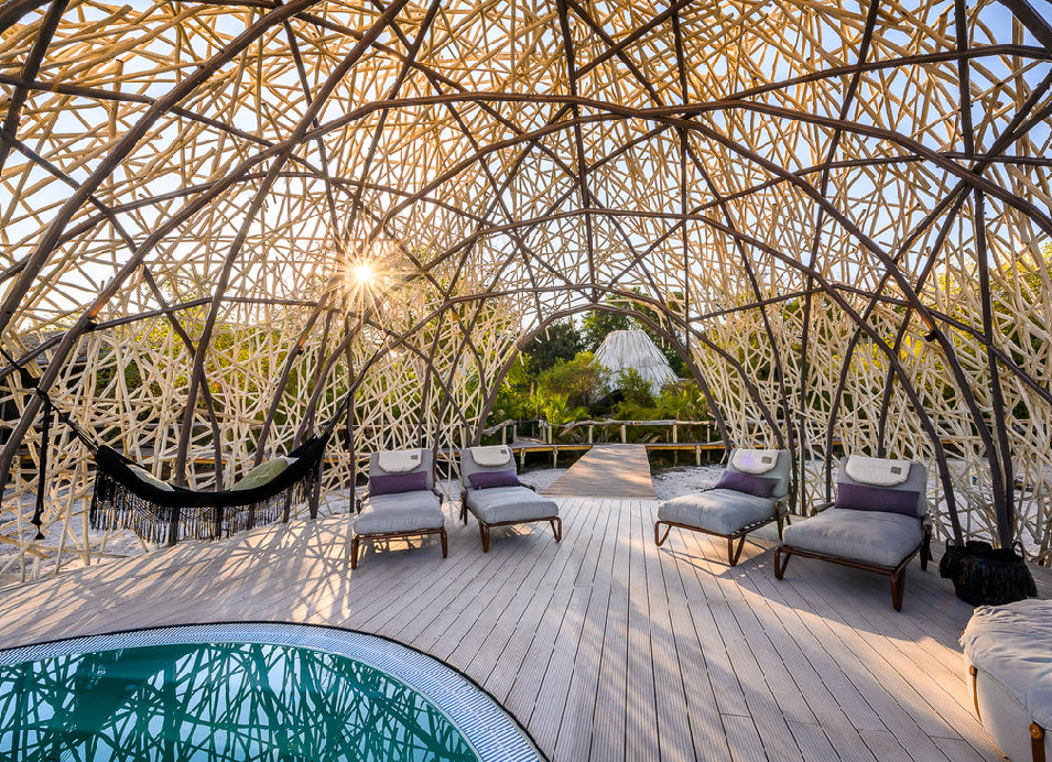 Outdoor lounge area at a Botswana safari camp, featuring a shaded woven branch structure, hammock, lounge chairs, wooden deck and a small pool surrounded by greenery.