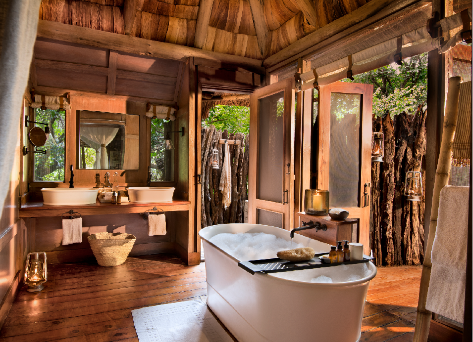 Luxury safari lodge bathroom with a freestanding bathtub, wooden vanity, and open-air views surrounded by natural wood and greenery in Tanzania.