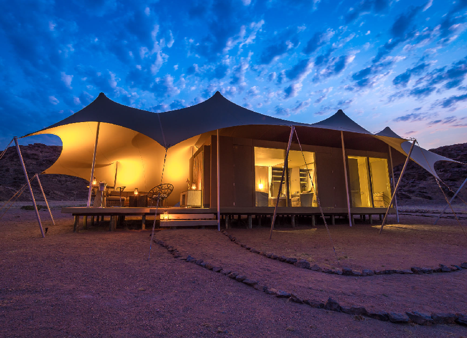 Luxury safari tent camp glowing at dusk in the rugged Namibian landscape beneath a colorful evening sky.