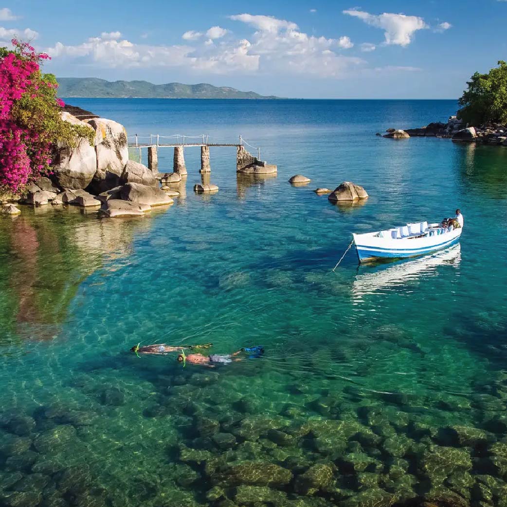 Scenic view of Lake Malawi with crystal-clear turquoise water, a small boat, swimmer, rocky shoreline, and mountains in the distance.