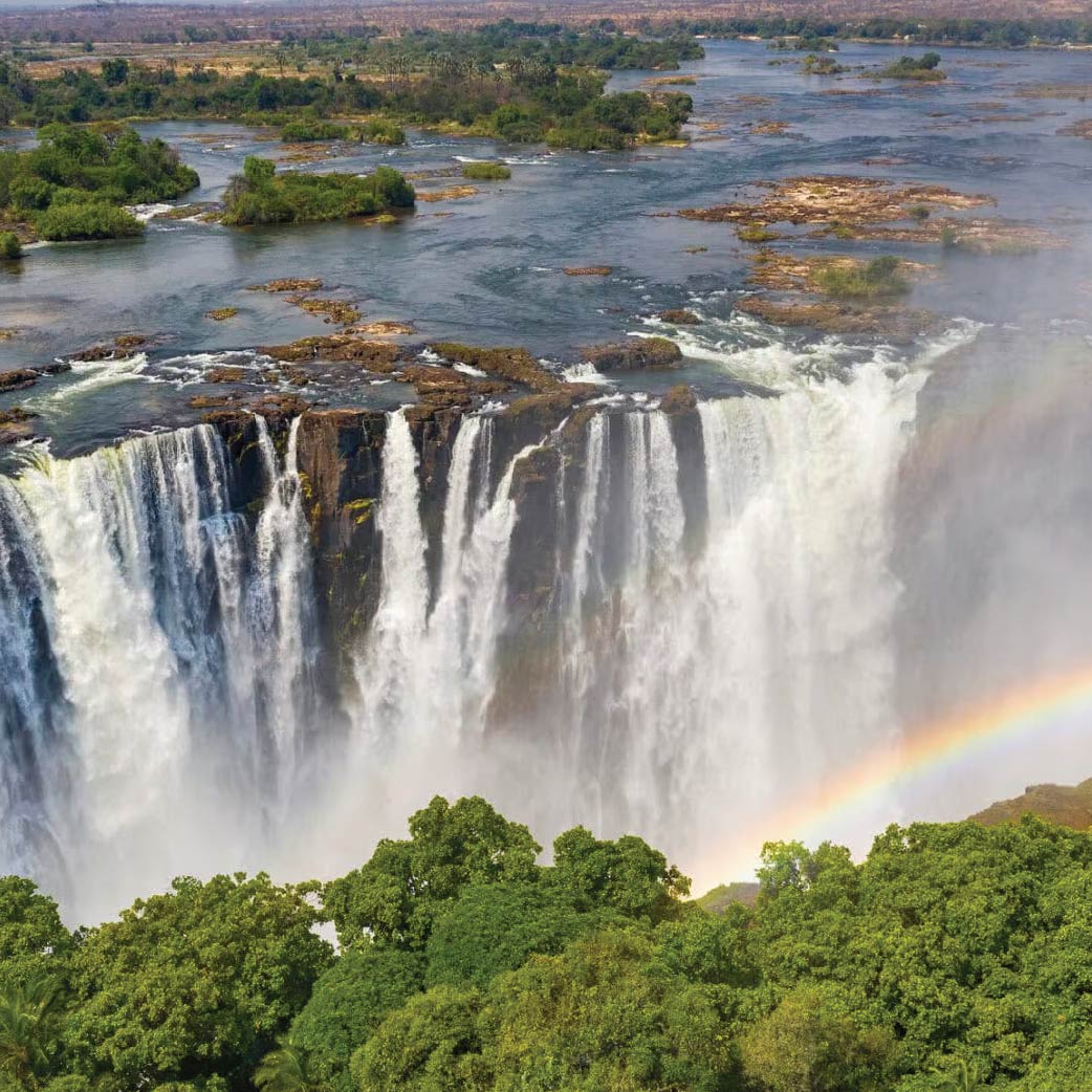 Aerial view of Victoria Falls in Zimbabwe with cascading waterfalls, lush greenery, and a rainbow.