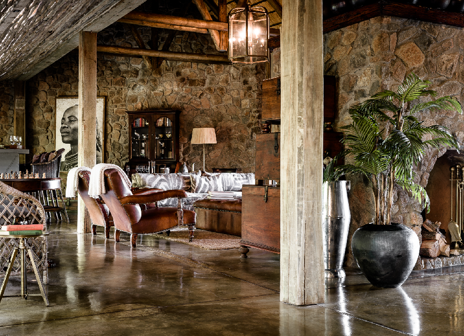 Elegant safari lodge lounge with stone walls, wooden beams, leather chairs, rustic furnishings, and tropical plants.