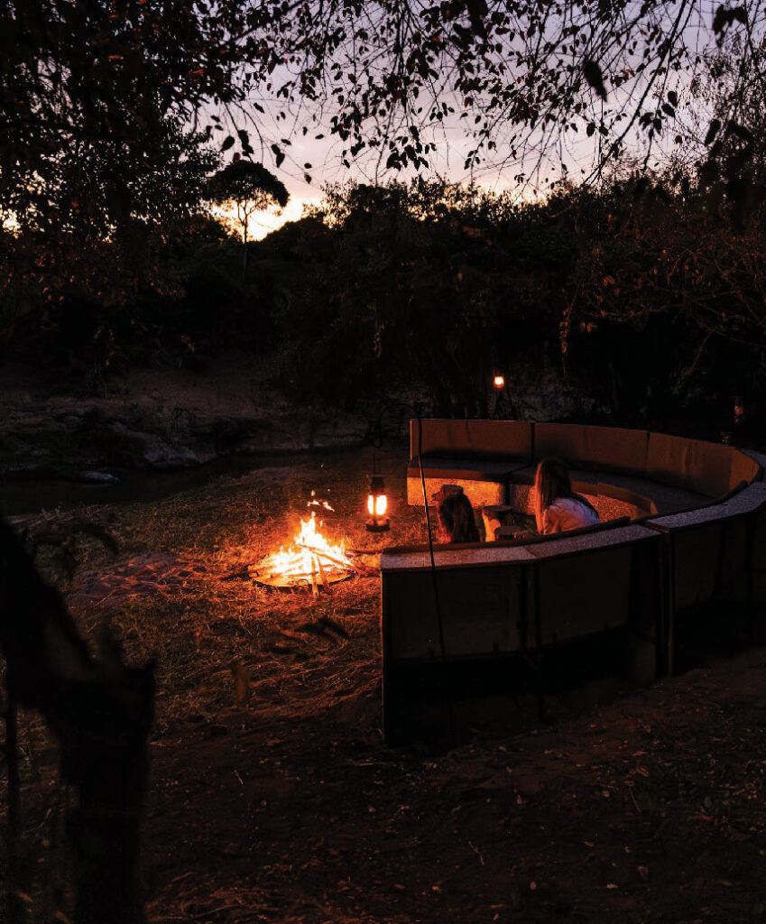 Outdoor safari campfire area with a fire burning beside a circular seating area surrounded by trees at dusk.