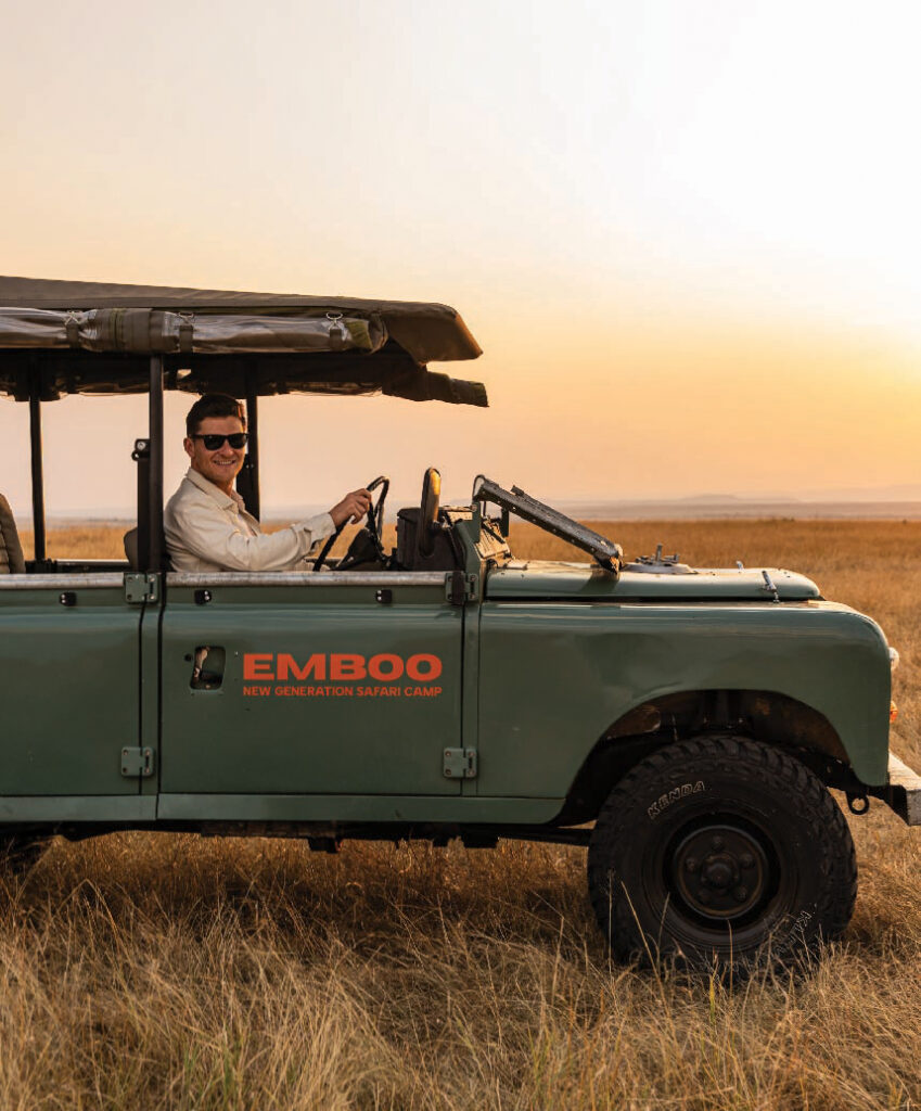 Man driving an Emboo safari Land Rover through dry grassland at sunset.