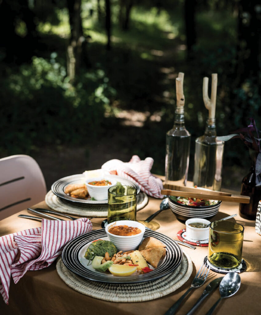 Outdoor picnic lunch setup with plates of food, drinks, and utensils on a wooden table surrounded by greenery.