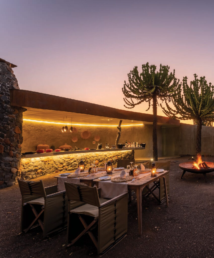 Outdoor safari dining area with a candlelit table, stone bar, and distinctive desert trees at sunset.