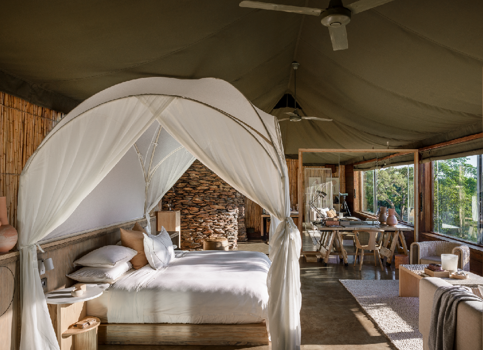 Safari tent interior with a four-poster bed, natural stone fireplace, dining area, and views of the African bush.