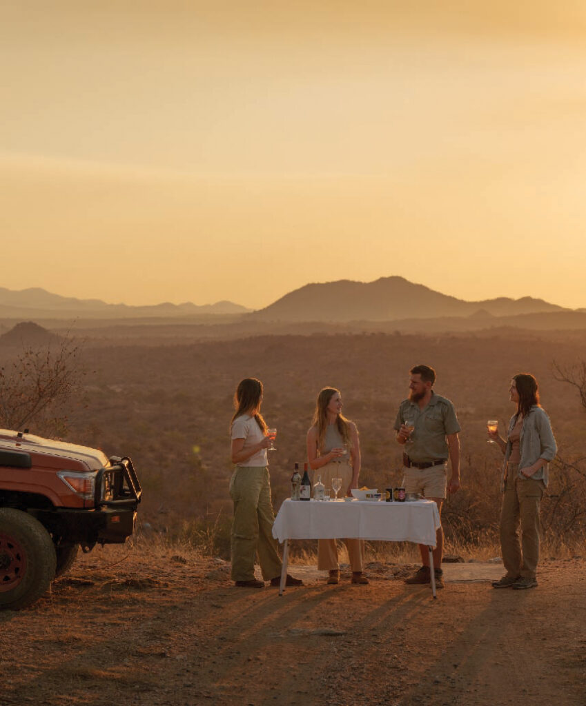 Four people enjoying a safari picnic beside a vehicle in an arid South African landscape at sunset, with mountains in the background.