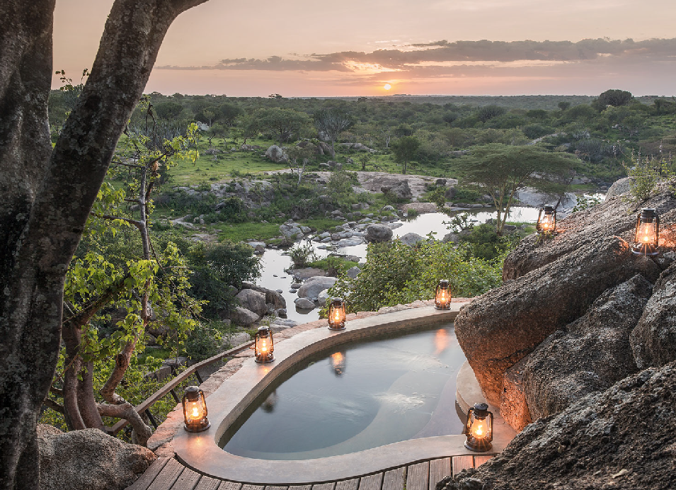 Luxury safari lodge pool overlooking a rocky landscape and lush African bush at sunset, with lanterns lining the pool.