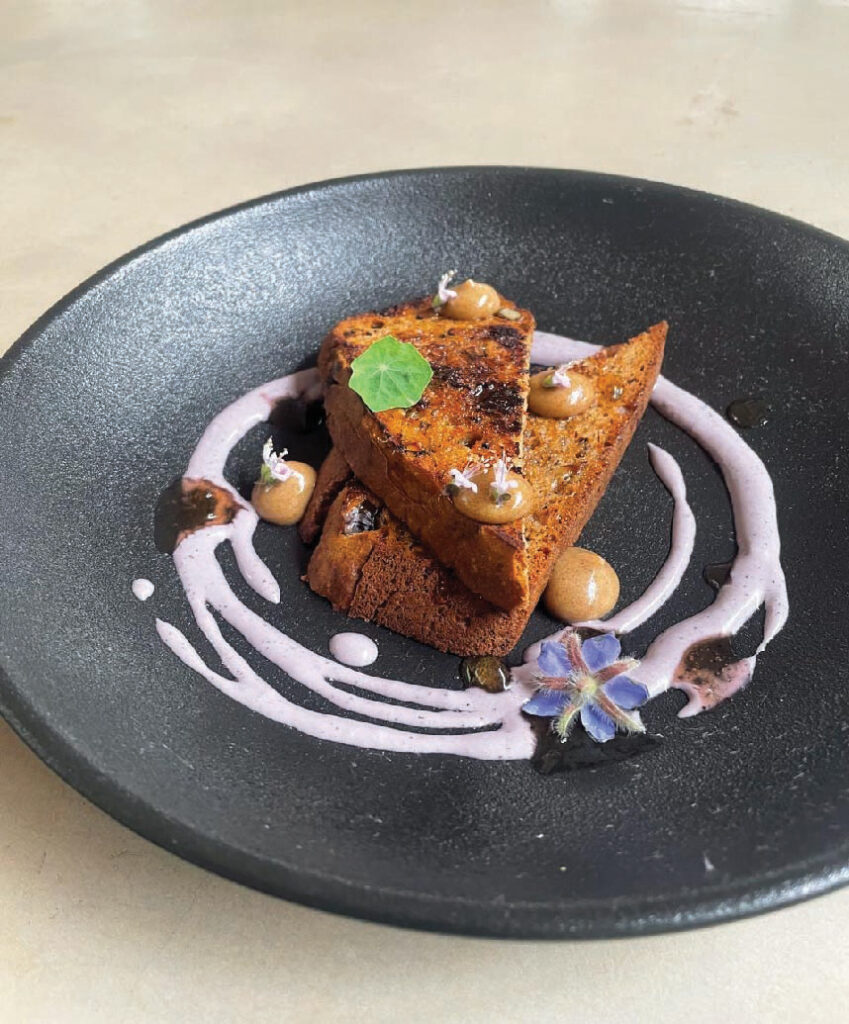 Artfully plated French toast with a crisp caramelized crust, creamy purple berry sauce, dollops of sauce, fresh mint, and an edible flower on a black plate.