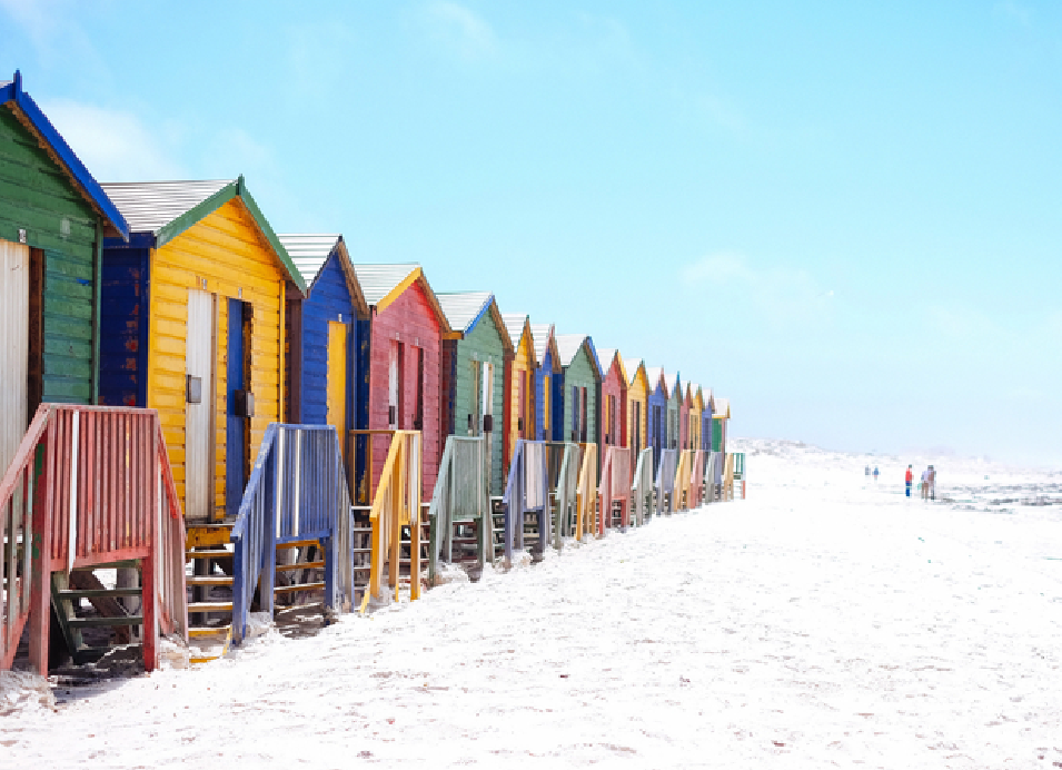 Colorful wooden beach huts lined along a white sandy beach with ocean waves in the background.