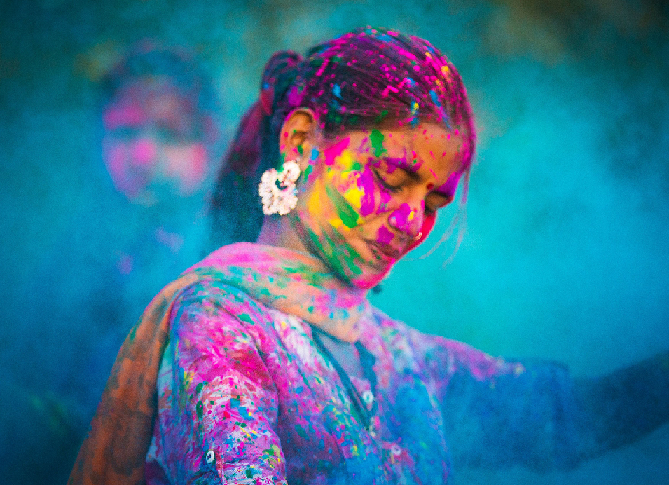 A woman covered in vibrant pink, yellow, green, and blue Holi colors, smiling and looking down during the colorful Indian festival.