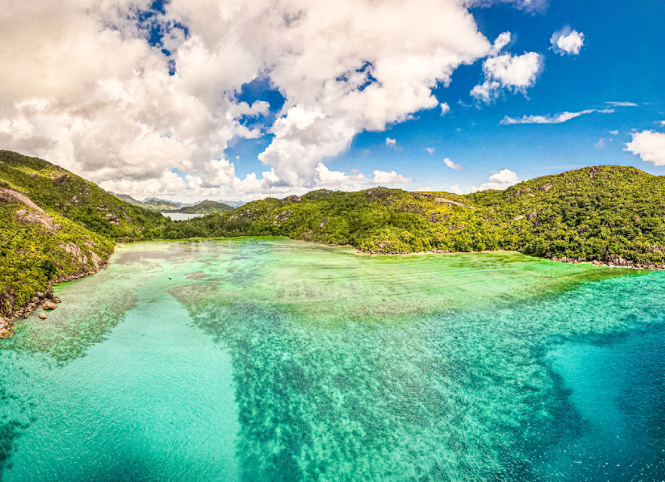 Tropical island lagoon with crystal-clear turquoise water, lush green hills, and a bright blue sky with clouds.
