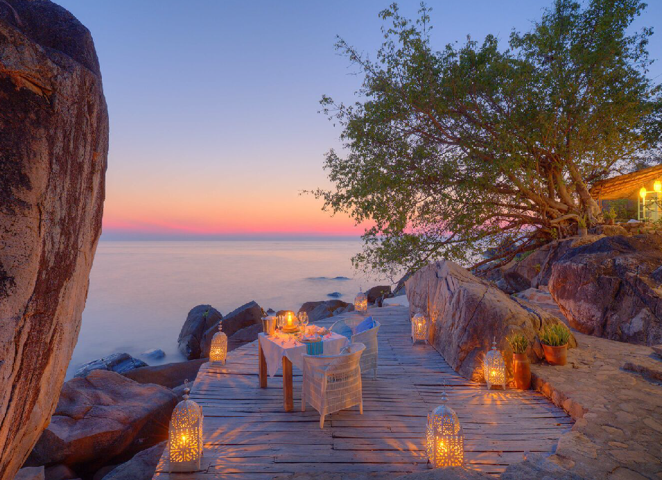Romantic lakeside dinner setup on a wooden boardwalk at sunset, with a table set for two, glowing lanterns, rocky shoreline, and a large tree overlooking the water.