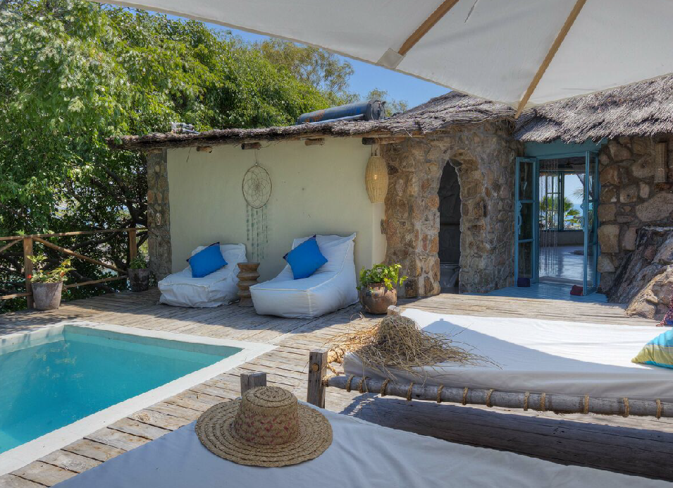 Private safari lodge pool area with white sun loungers, blue cushions, thatched-roof stone accommodation, and a shaded outdoor daybed.