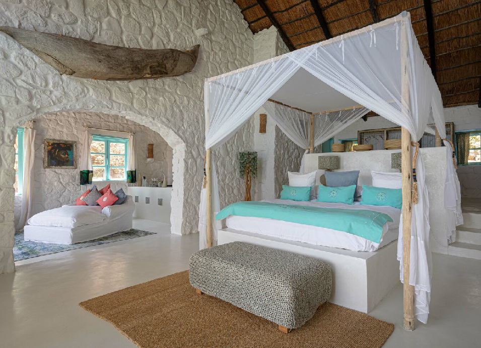 Luxury safari lodge bedroom with a four-poster canopy bed, white stone walls, turquoise bedding, and natural wood accents.