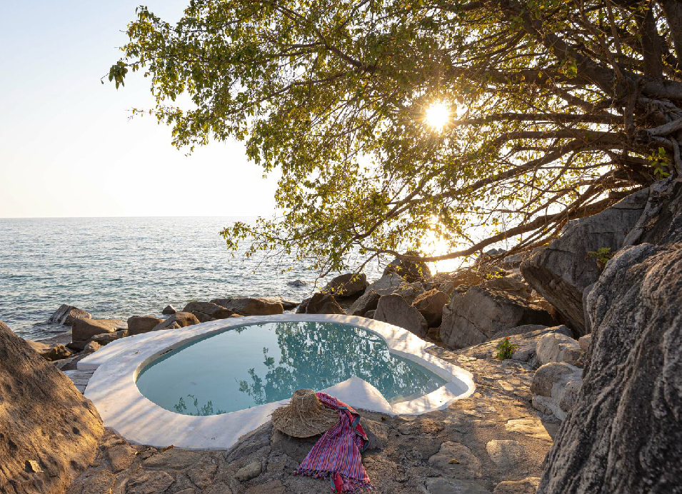 Secluded infinity pool overlooking the ocean, surrounded by rocky shoreline and a large tree at sunset.