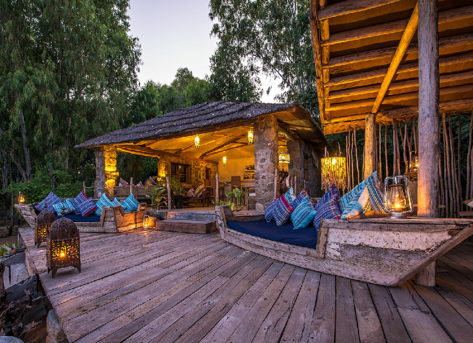 Kaya Mawa lodge in Malawi with a rustic wooden deck, thatched buildings, colorful cushions, and warm evening lighting surrounded by trees.