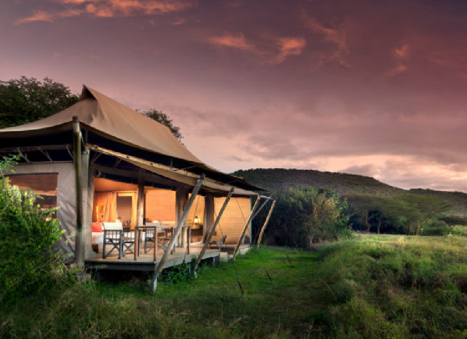 Luxury safari tent lodge surrounded by lush grassland and trees at sunset, with a private outdoor veranda.