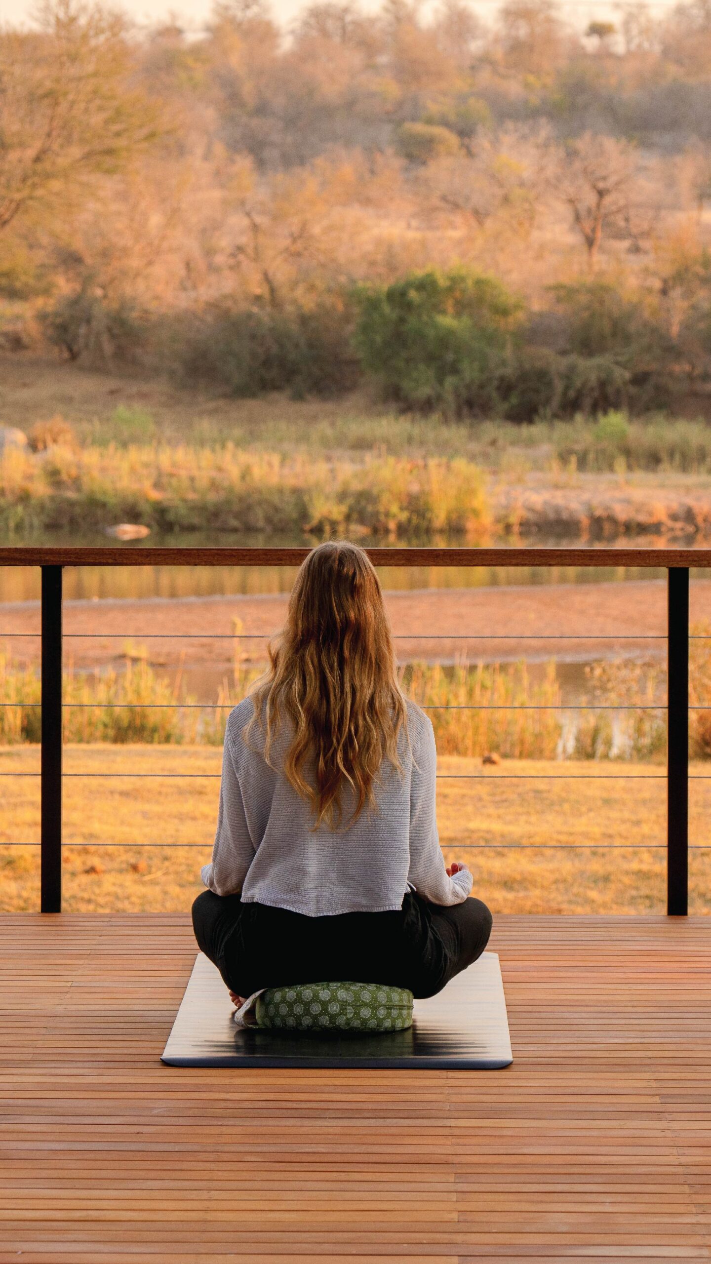 Woman meditating on a yoga mat on a wooden deck overlooking a peaceful wildlife landscape in South Africa.