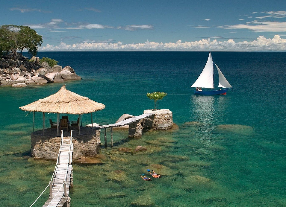 MalawiIsland beach in Malawi with a thatched-roof hut over turquoise water, a wooden pier, swimmers, and a sailboat on Lake Malawi.
