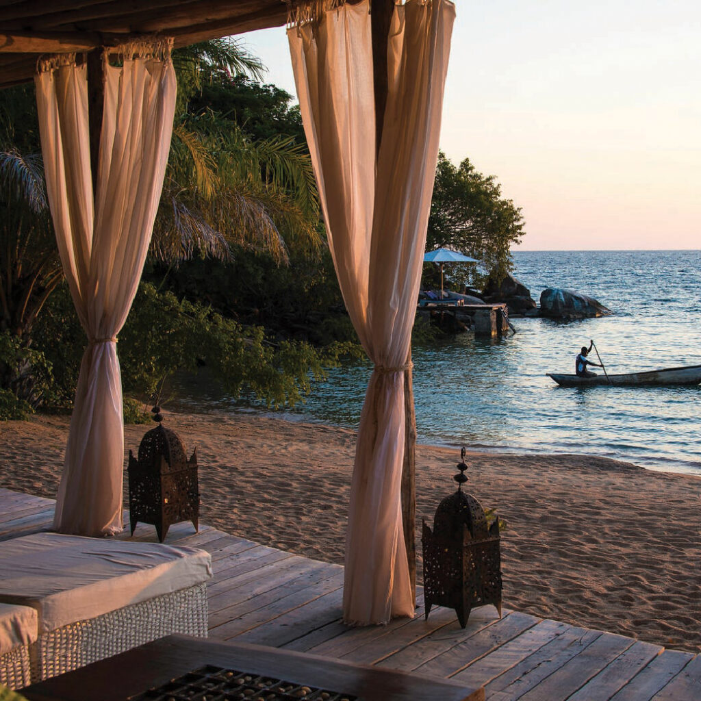 Beachfront safari lodge with a shaded canopy, overlooking the ocean and a small boat at sunset in Malawi.