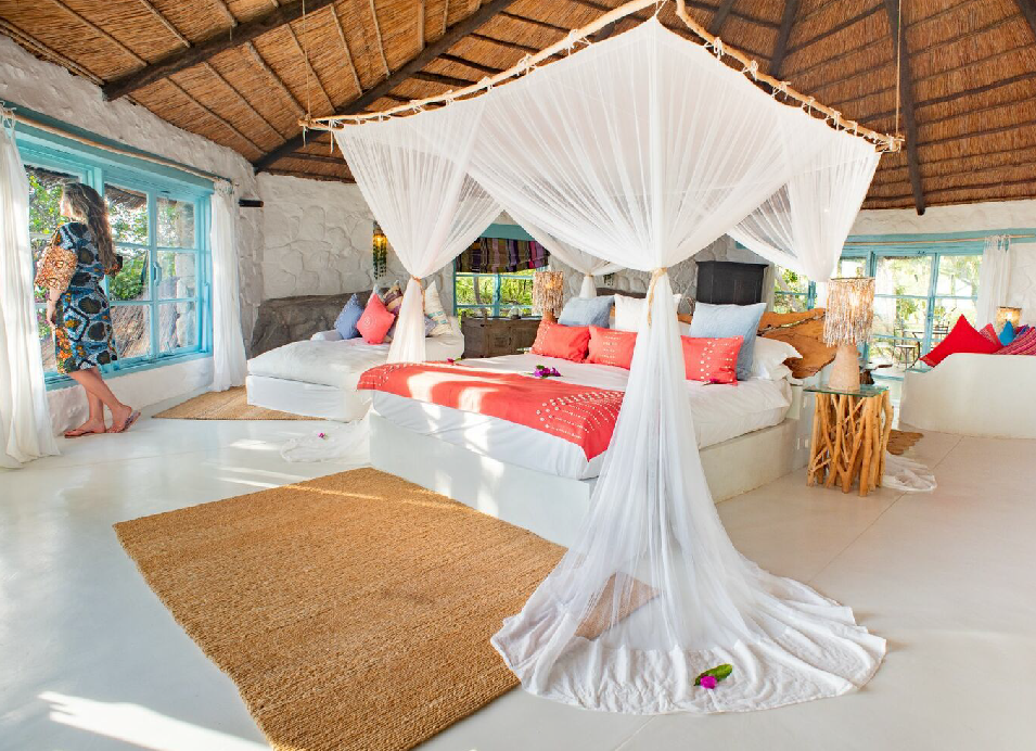 Bright tropical lodge bedroom with a four-poster bed, white mosquito net, colorful cushions, and thatched roof