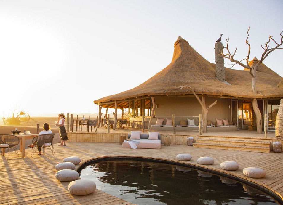 Safari lodge with thatched roofs, a wooden deck, outdoor seating, and a pool overlooking the Namibian desert at sunset.