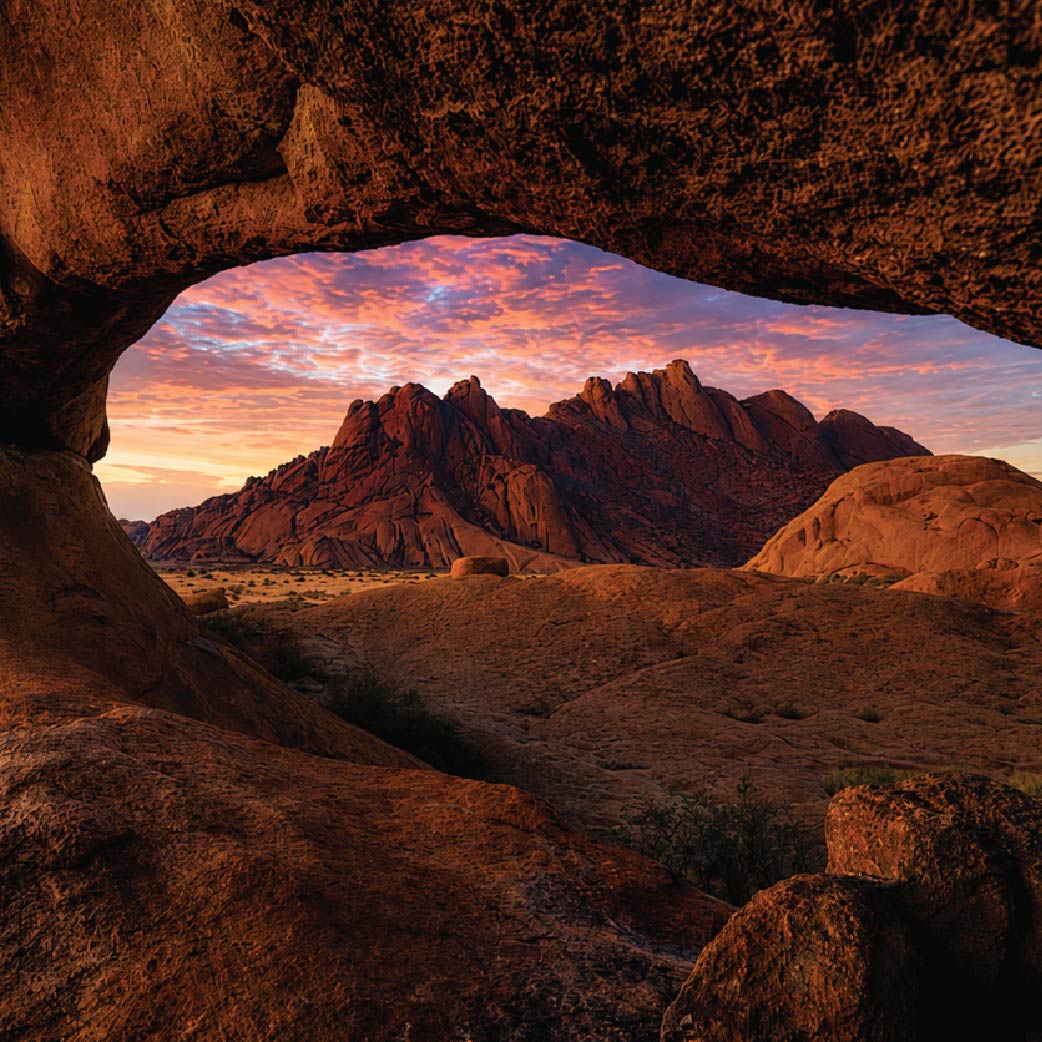 Landscape in Namibia framed by a natural rock arch, with rugged peaks beneath a colorful sunset sky.