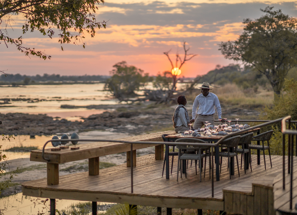 Guests enjoying an outdoor dining experience on a riverside deck at sunset in Zambia.