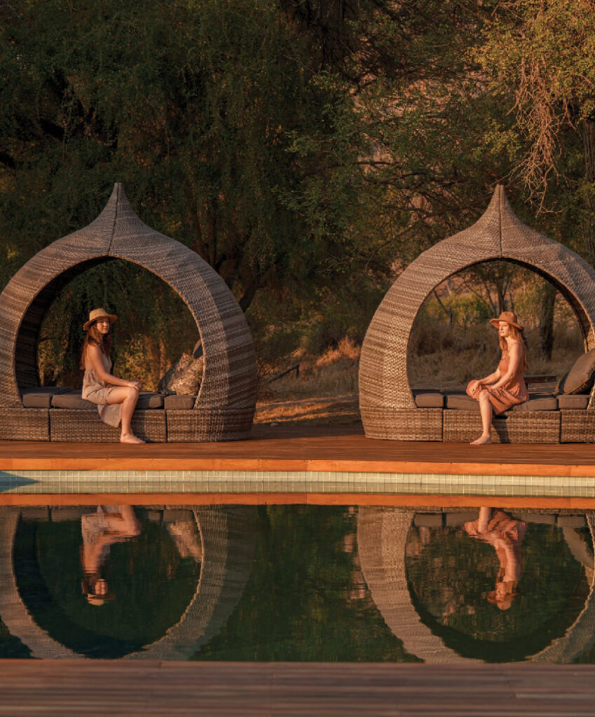 Two women in hats sit in woven egg-shaped chairs beside a swimming pool at a safari lodge in South Africa, surrounded by trees, with their reflections visible in the water.