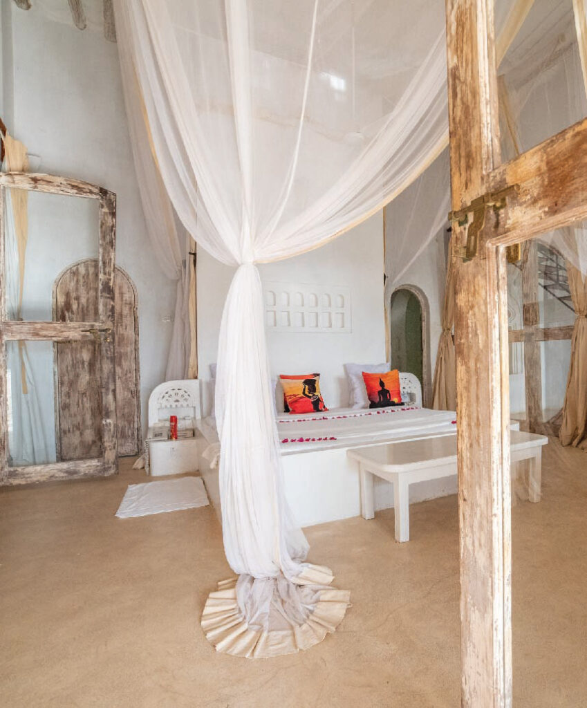 Bright, elegant safari lodge room with a white canopy bed, rustic wooden beams, white furnishings, and colorful decorative cushions.