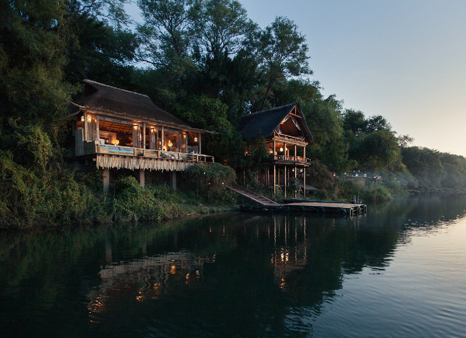 Lodge overlooking the Zambezi River at sunset, surrounded by trees and reflecting in the calm water.