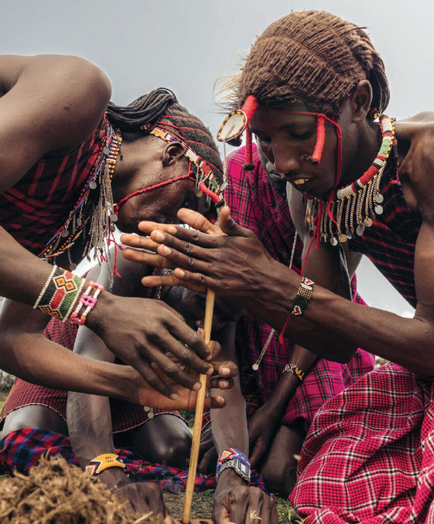 Maasai people in traditional red clothing and beaded jewelry performing a cultural ceremony, with one holding a wooden stick.
