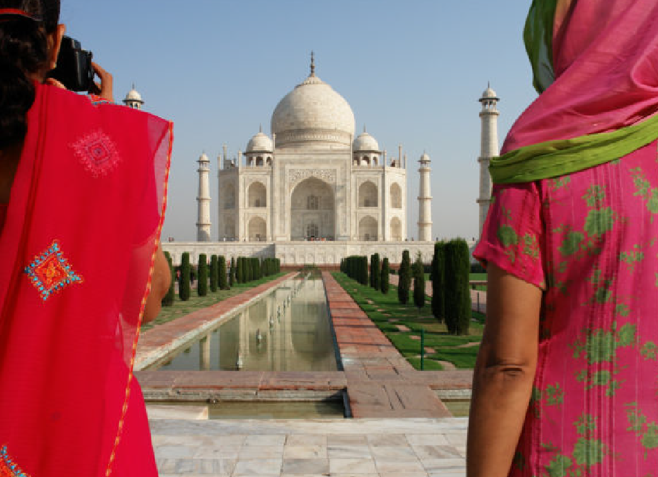 The Taj Mahal in Agra, India, viewed through two women in colorful traditional clothing, with the reflecting pool and gardens in the foreground.