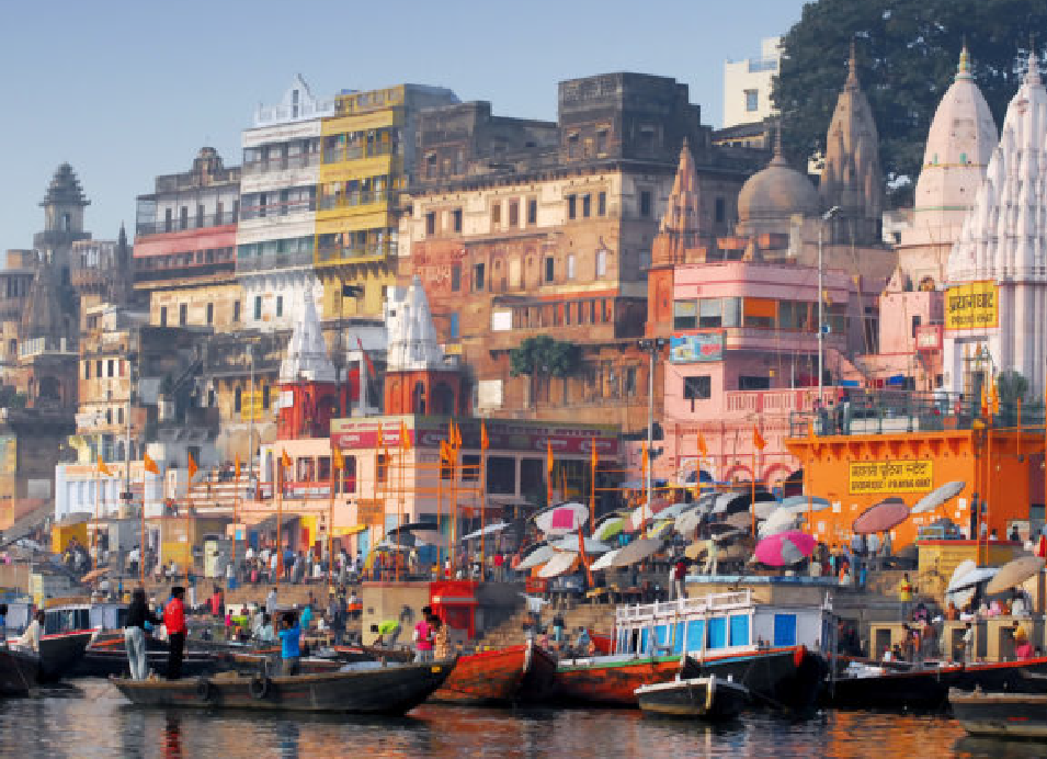 Varanasi ghats along the Ganges River in India, with colorful boats, umbrellas, historic buildings, and Hindu temples lining the waterfront.