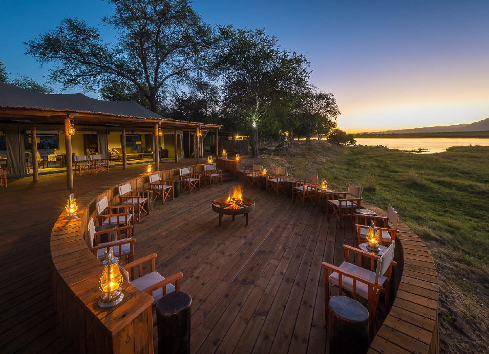 Luxury safari lodge accommodation with a wooden deck, outdoor seating, lanterns, and a fire pit overlooking a river at sunset