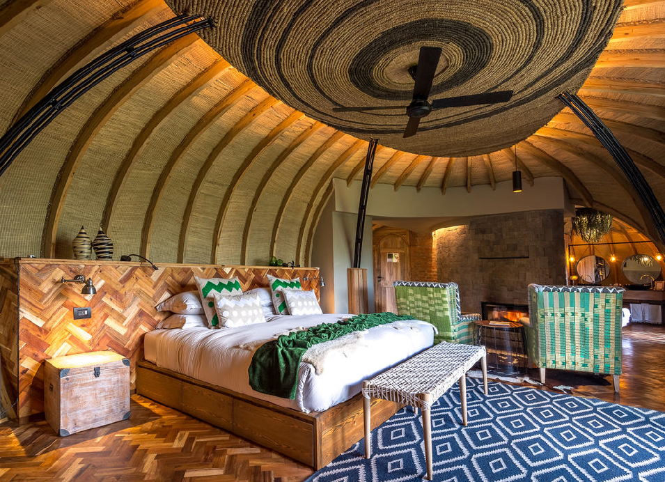 Luxury safari lodge bedroom with a king-size bed, woven décor, wooden floors, and a large round ceiling fan.