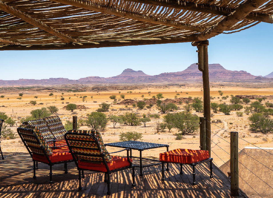 Rustic safari seating area under a thatched shade structure overlooking the desert landscape and mountains of Namibia.