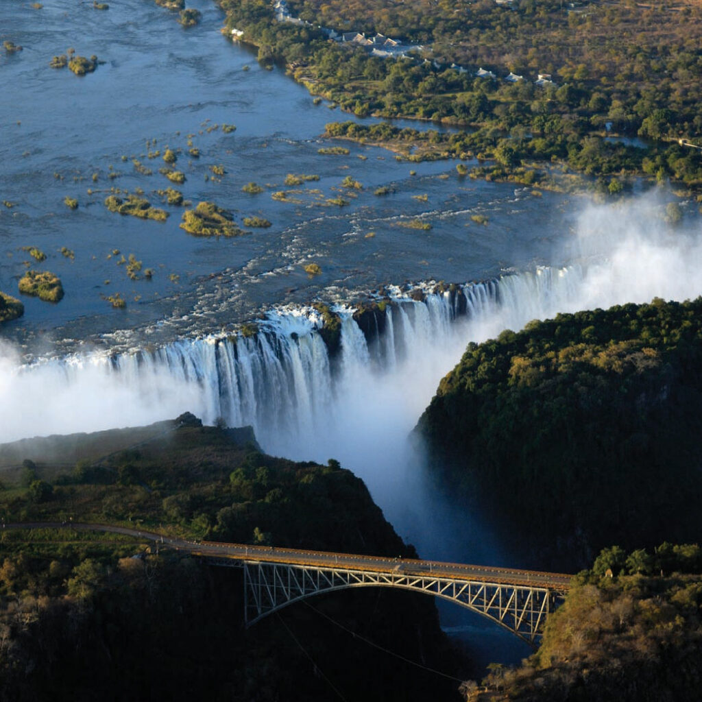Aerial view of Victoria Falls in Zambia, with the Zambezi River flowing over the falls and a historic bridge crossing the gorge below.